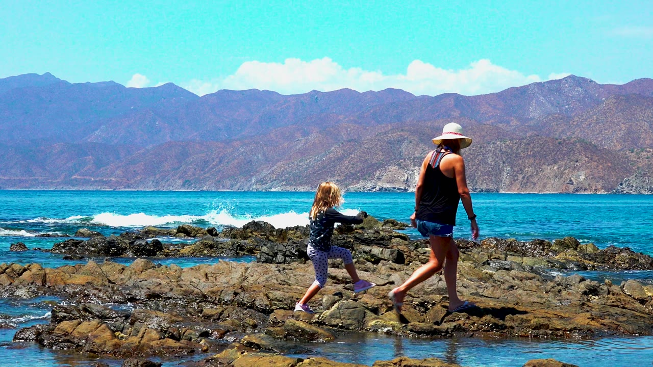 A woman in a white hat and a young child walk across dark rocks on the El Sargento coastline, with blue ocean, white surf, and mountains behind them.