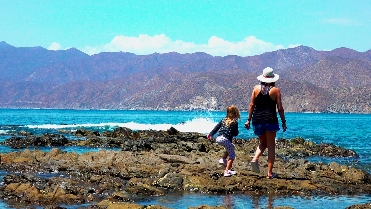 Child steps ahead while woman follows across shoreline rocks.