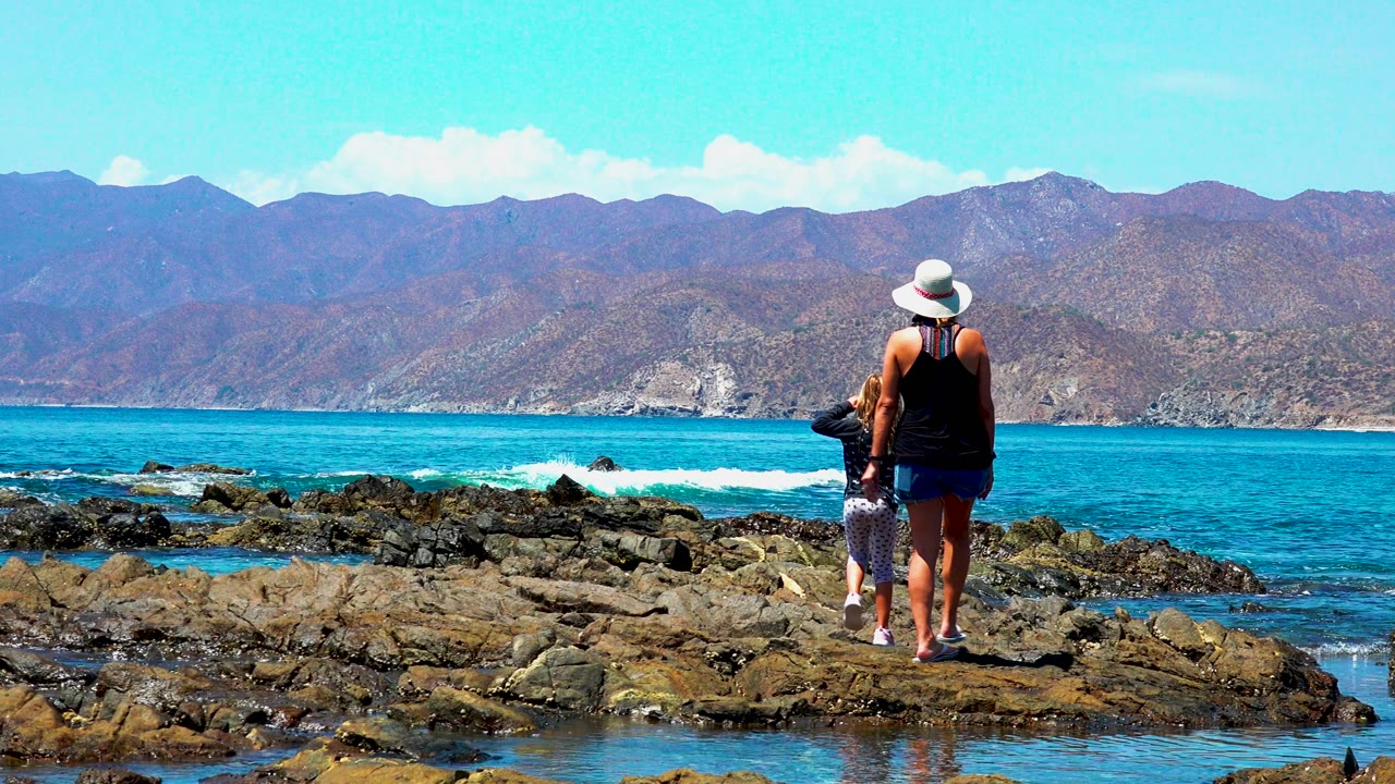 The woman and child stand near the right side of the rocky El Sargento shoreline as white surf breaks across the blue water and mountains span the horizon.