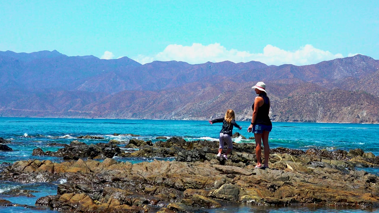 At El Sargento, Mexico, a woman in a white hat stands on shoreline rocks while a young child faces the blue ocean and distant mountains.