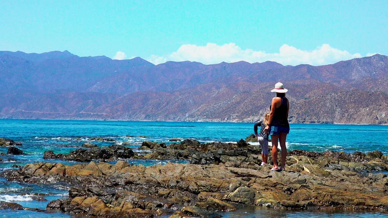 Woman and child paused on dark rocks with ocean and mountains beyond.