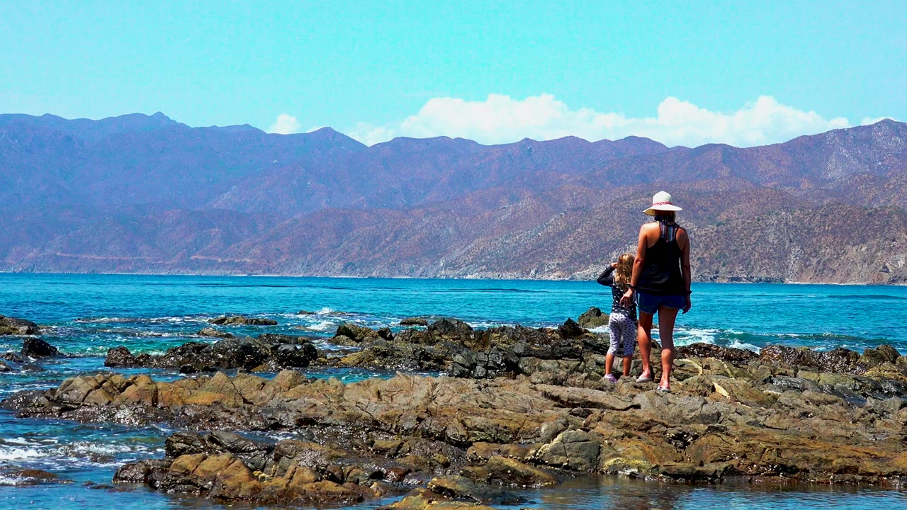 At El Sargento, Mexico, the woman and child appear near the right edge of the frame on uneven rocks, with a wide band of blue sea and mountains stretching behind them.