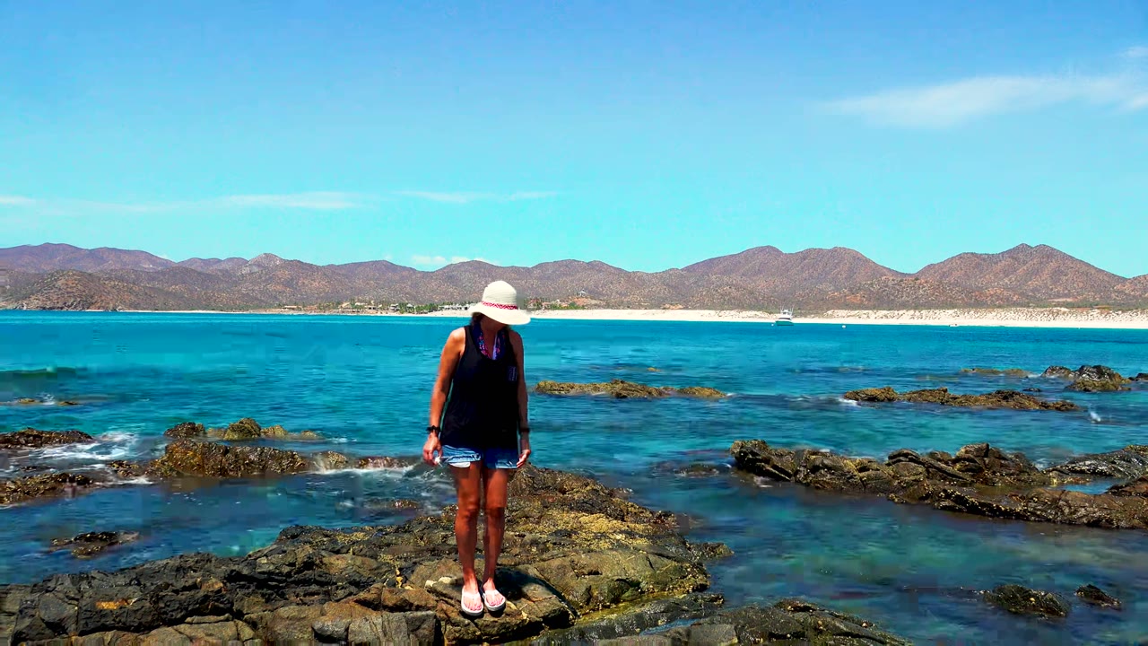 At El Sargento and La Ventana, Mexico, a woman in a white hat stands near the left side of dark shoreline rocks with blue water, pale sand, and low mountains behind her.