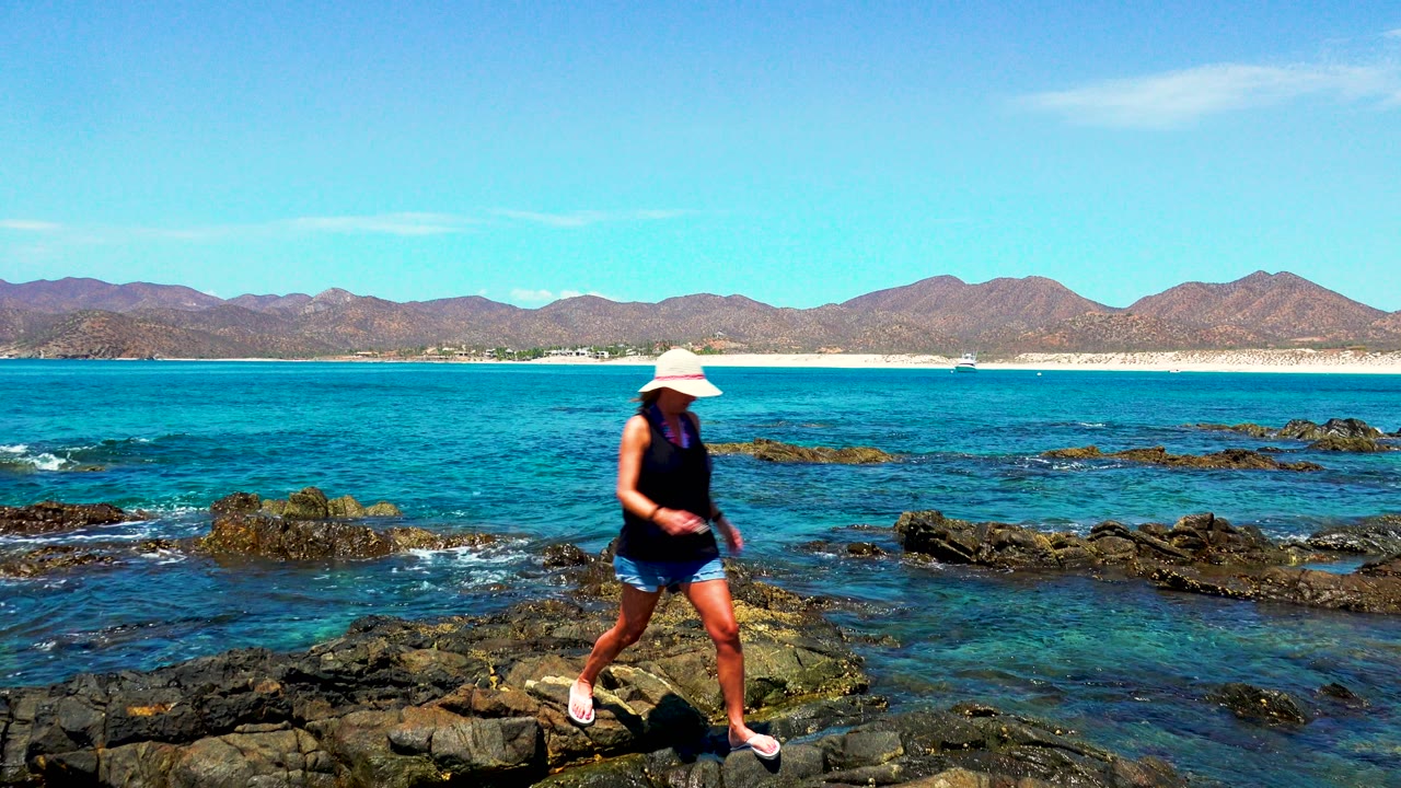 The woman walks left across uneven dark rocks at El Sargento and La Ventana, Mexico, with shallow blue water around the rock formations and a mountain ridge across the horizon.