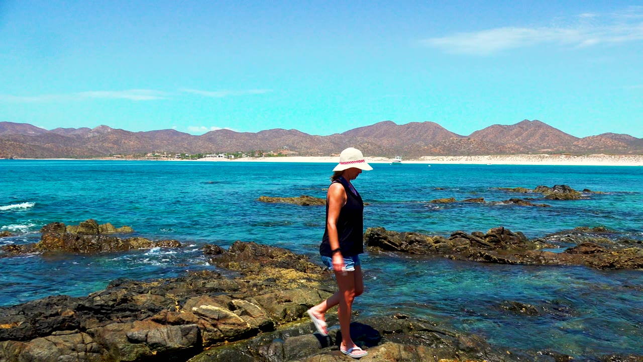 Near the right edge of the frame, the woman continues along the rocky shore in El Sargento and La Ventana, Mexico, while small waves break around dark rocks in the blue water.