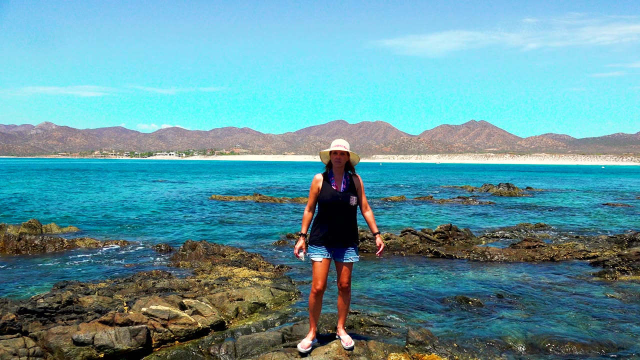 Woman facing camera on rocky shore with blue ocean and mountains in back.