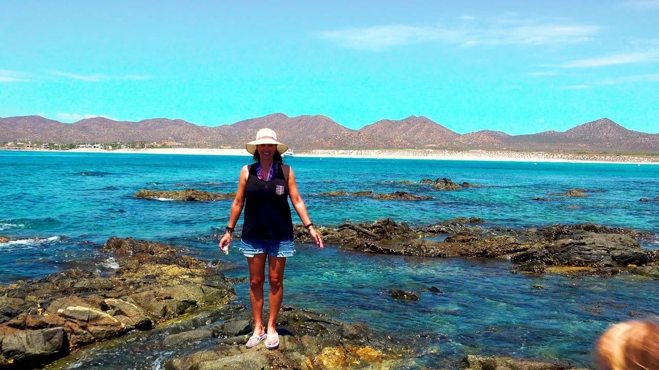 Woman in a sun hat stands alone on the rocky shore in El Sargento, Mexico, with blue water, low waves, sandy shoreline, and distant mountains behind her.