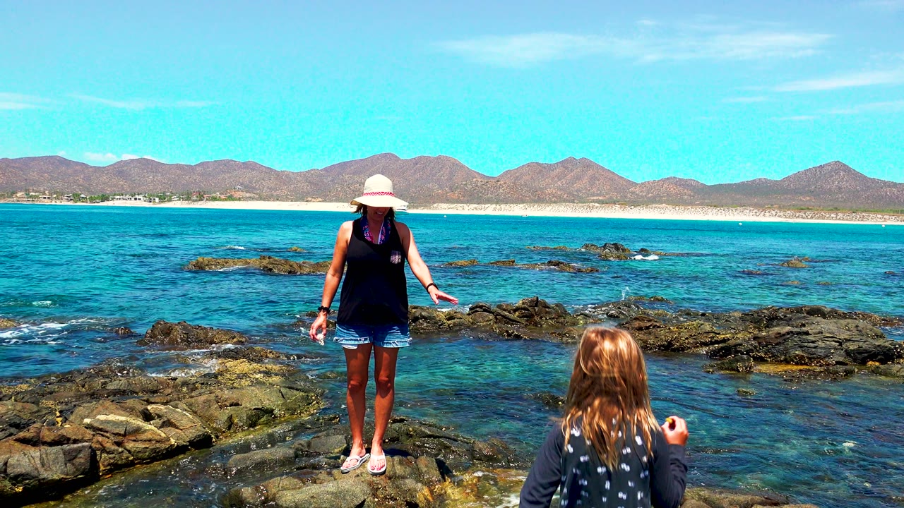 Woman on rocky shoreline as a child appears at the bottom edge of frame.