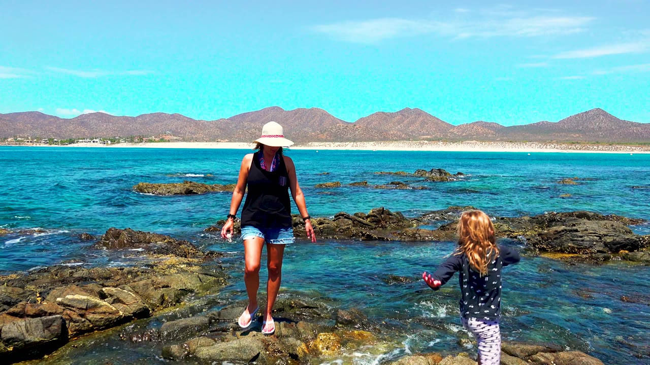 A child in a dark patterned top approaches the woman across rocks in El Sargento, Mexico, with shallow turquoise water, dark rock outcrops, and mountains in the background.