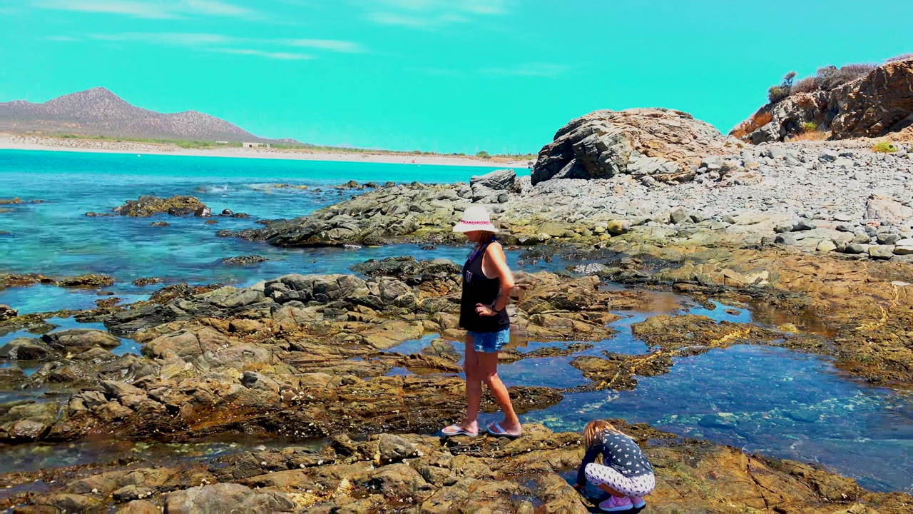 At 0:00 in El Sargento, Mexico, an adult in a sun hat stands on dark coastal rocks while a child crouches near tide pools with blue water and distant mountains behind them.