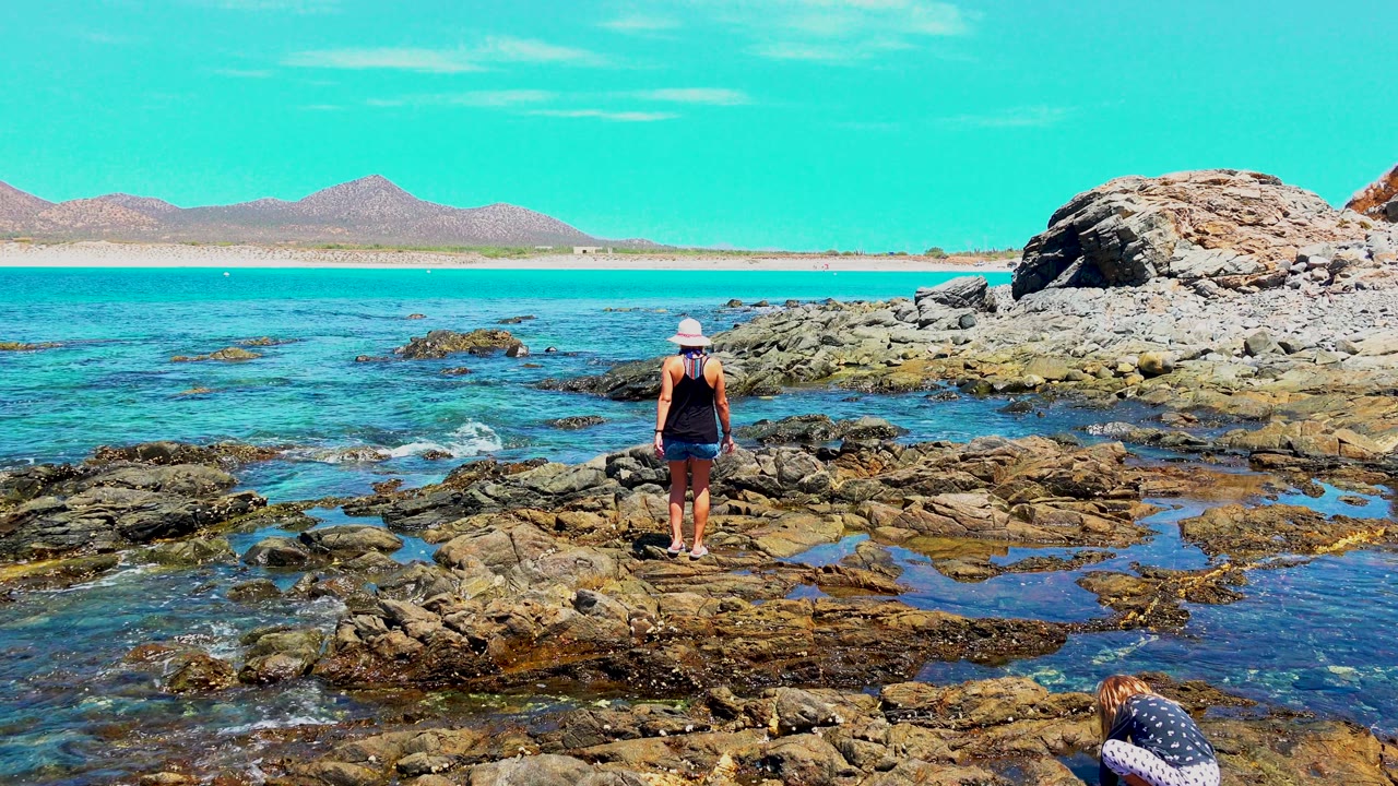 At El Sargento, Mexico, a woman in a white hat stands on wet coastal rocks beside turquoise water, with a crouching person partly visible near the lower edge and hills across the horizon.