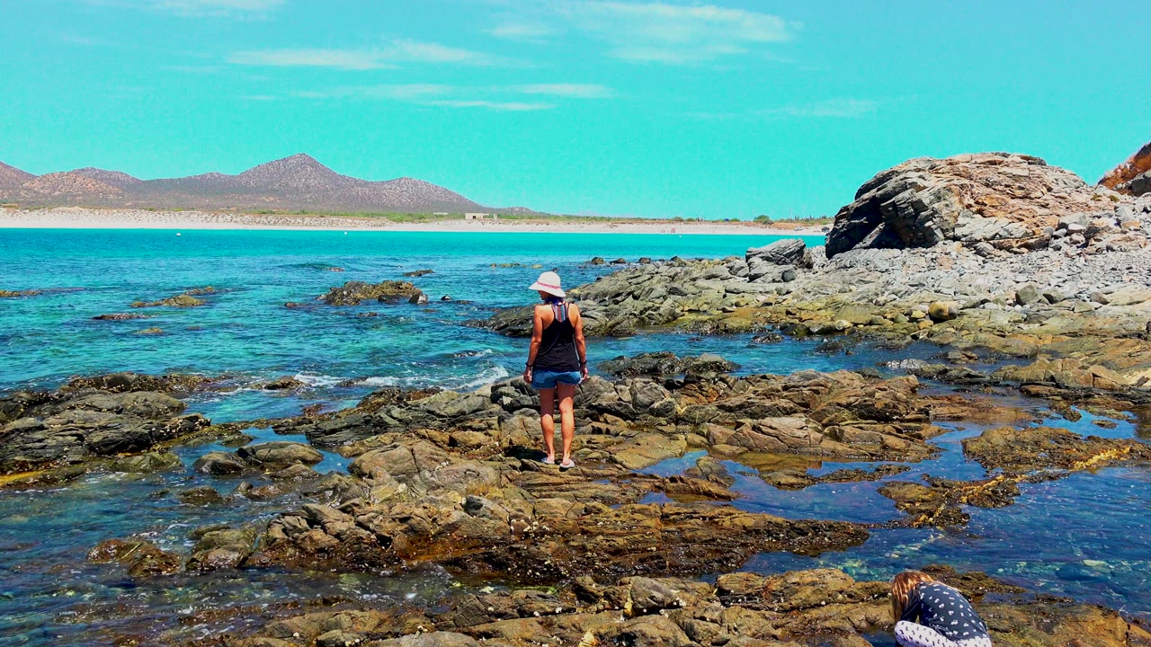 Woman on rocky coastal terrain facing clear blue water.