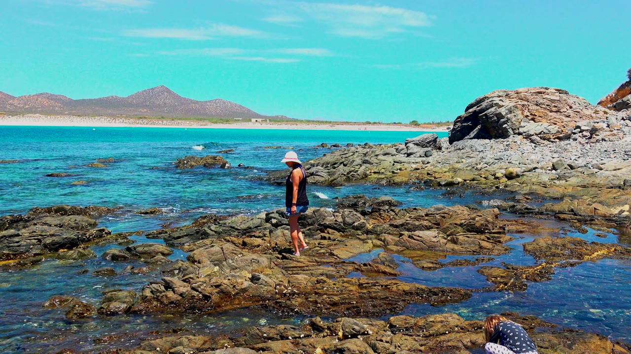 At El Sargento, Mexico, the woman has moved to the right across the uneven rocks, with turquoise water, scattered offshore rocks, a large outcrop, and distant hills behind her.