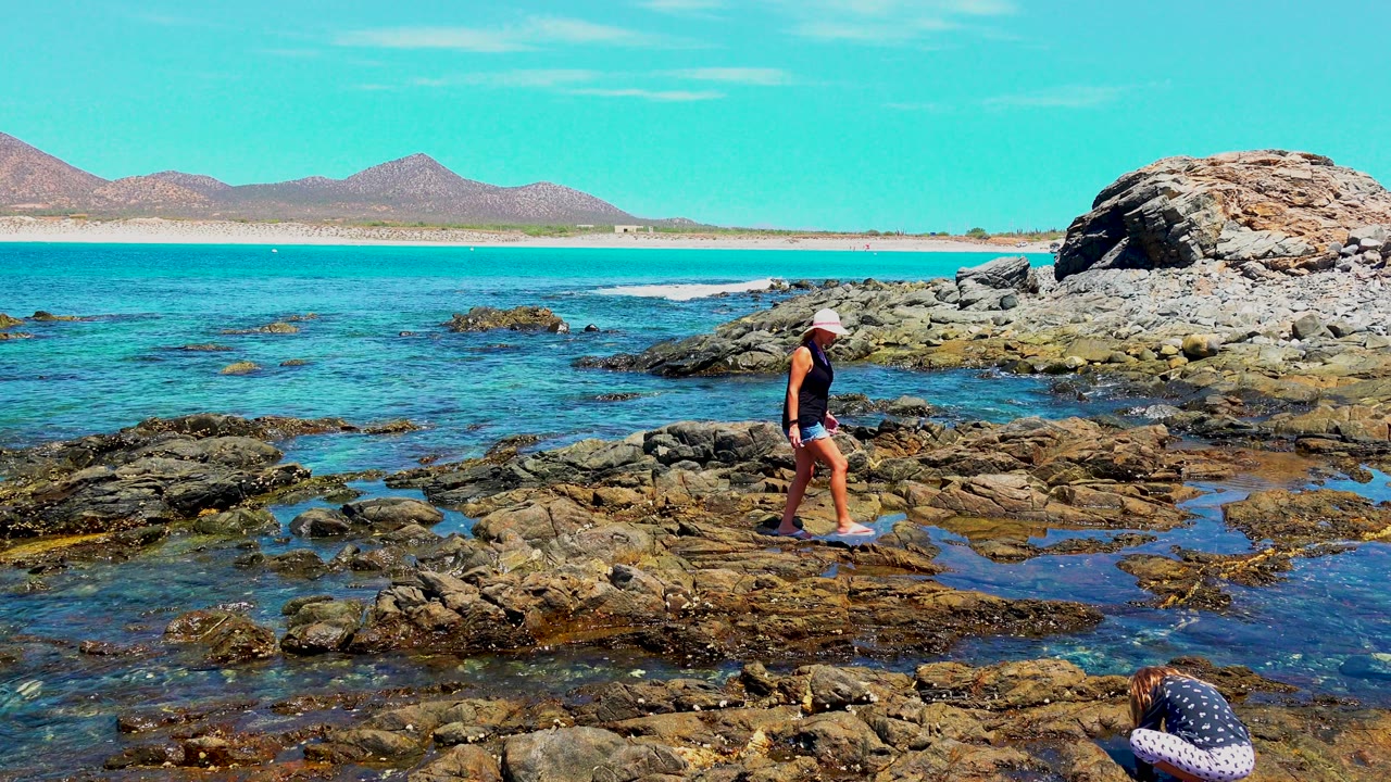 Adult in a hat walks left to right across rocky tide pools in El Sargento, Mexico, with a crouching child partly visible at the lower edge and distant hills across the water.