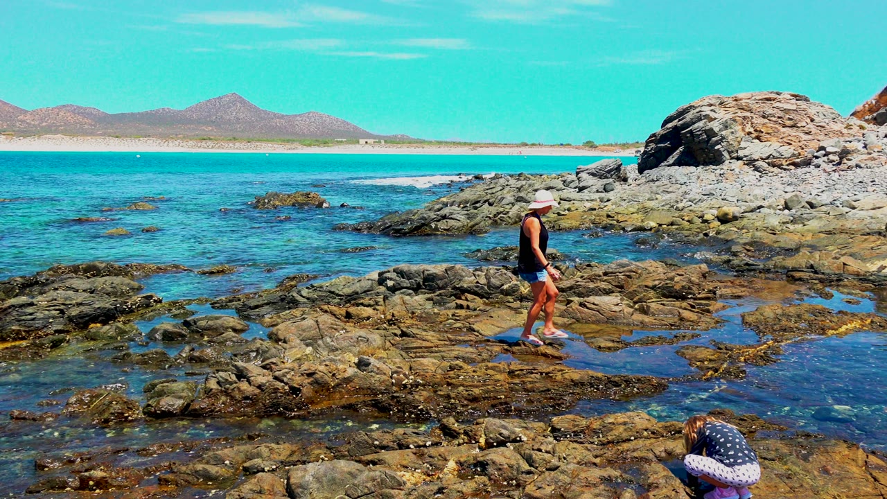 Adult continues across the wet rock shelf in El Sargento, Mexico, with blue tide pools in the foreground, turquoise water behind, and a child crouched low near the bottom edge.