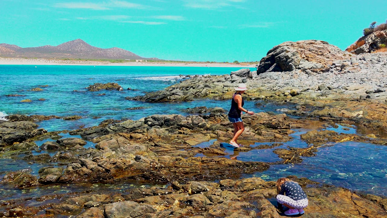 Adult in casual beach clothing moves farther right across the rocky El Sargento shoreline, with small waves, clear water, a sandy strip, and distant hills visible behind.