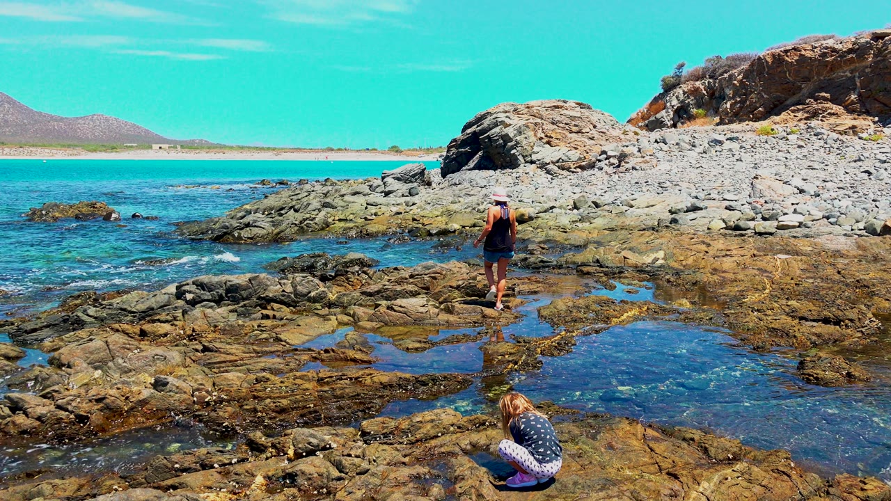 At El Sargento, Mexico, a child crouches beside shallow tide pools in the lower foreground while an adult stands on wet rocks near clear blue water and a pebbled shore.
