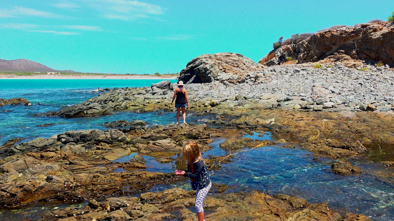 At El Sargento, Mexico, a child stands in the right foreground by shallow tide pools while an adult stands farther back on the rocky shore near turquoise water.