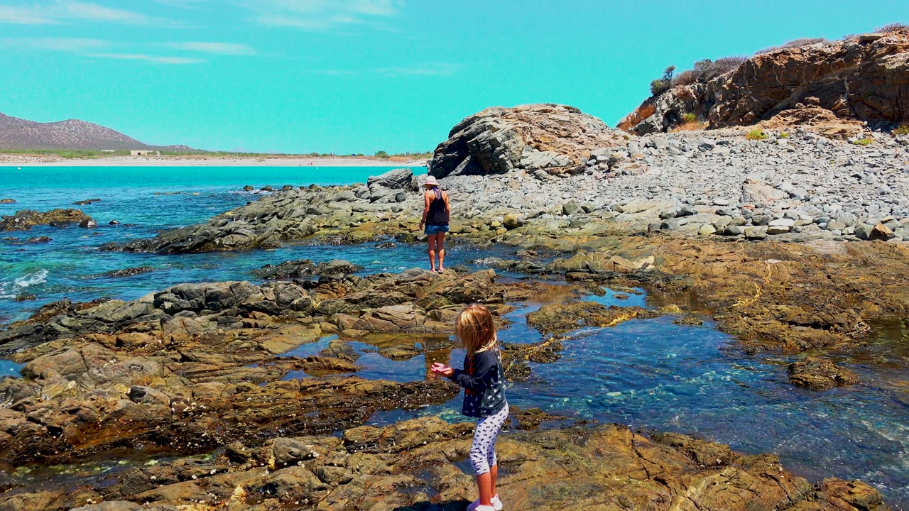 At El Sargento, Mexico, the child stands closer to the lower right edge beside a reflective pool while the adult faces the water from the center of the rocky shoreline.