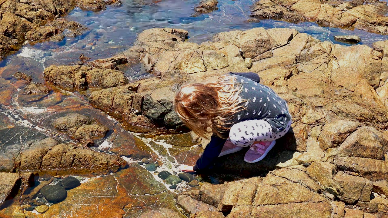 At 0:00, a young girl crouches low on sunlit rocks beside shallow clear water on the El Sargento, Mexico shoreline.