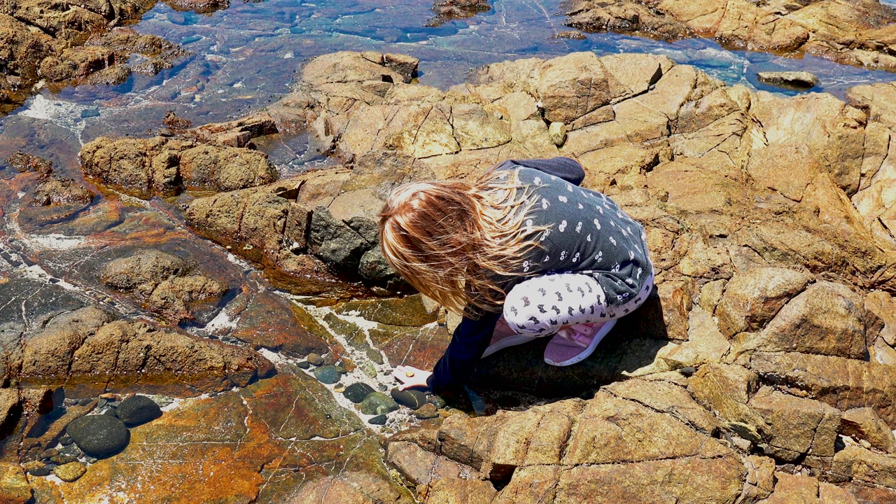 The child stays low over orange-stained and tan shoreline rocks in El Sargento, Mexico, with rippled blue tide-pool water behind her.