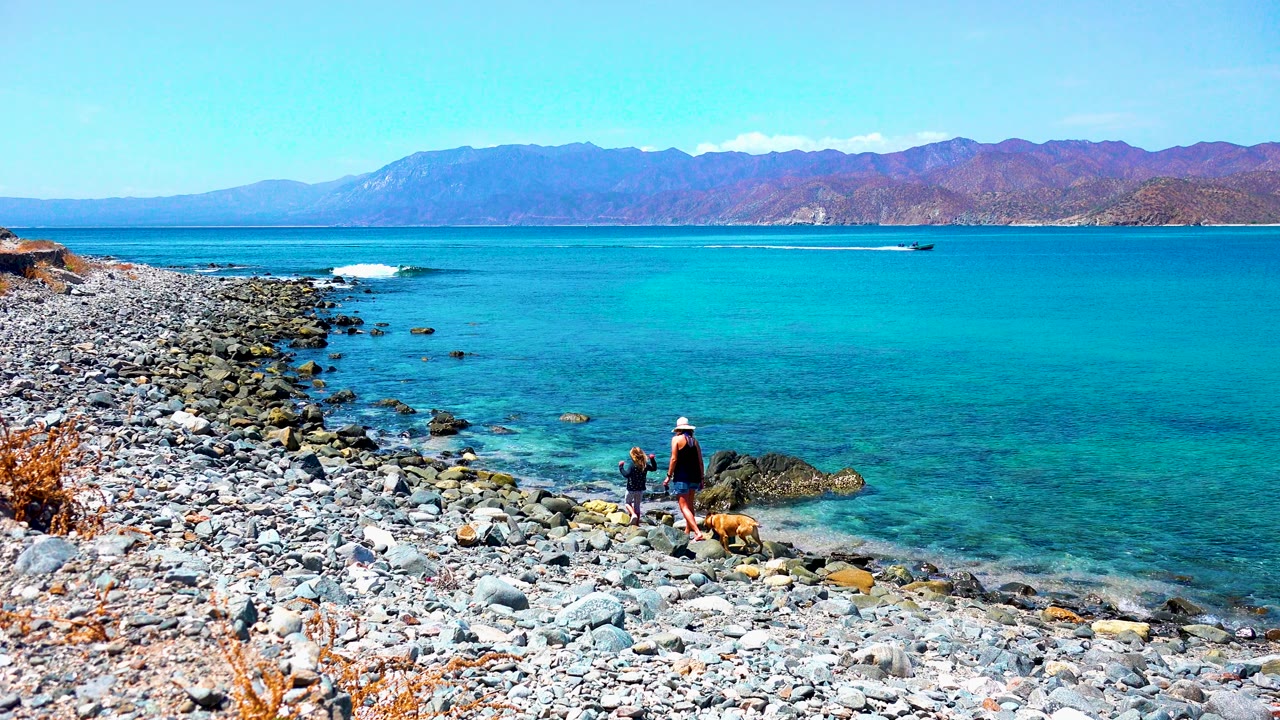 At El Sargento, Mexico, two people and a dog stand near rocks at the waterline, with turquoise water, distant mountains, and dry grasses along the rocky foreground.