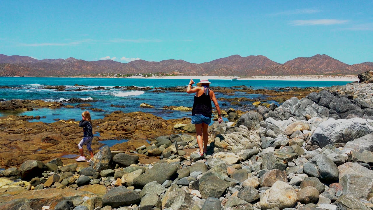 Adult in a sun hat and a child walk on a rocky shoreline in El Sargento, Mexico, with blue water, low waves, and mountains across the horizon.
