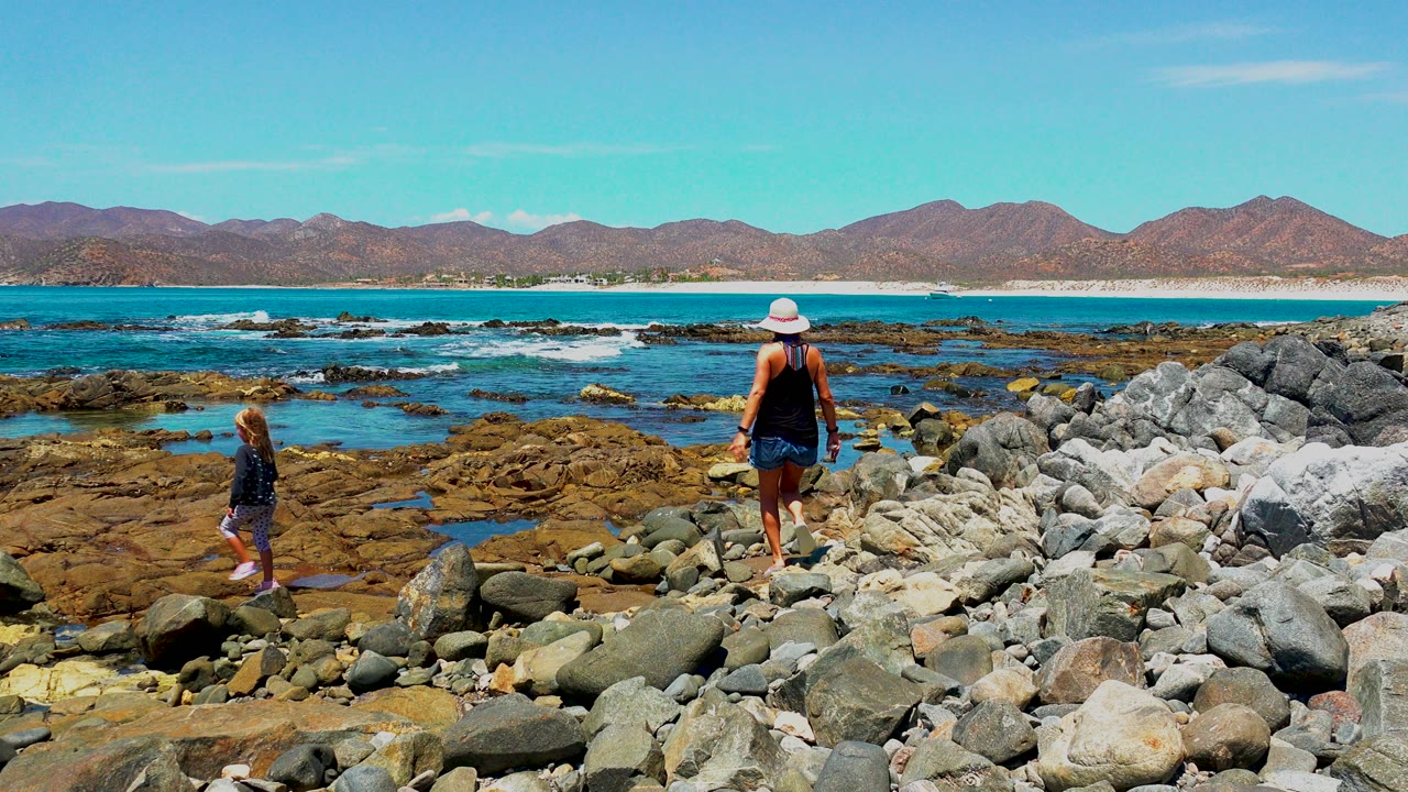 Child steps across rocks while adult stands farther back near the water.