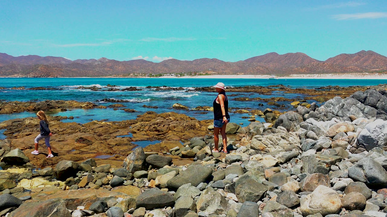 Adult in a light hat stands on pale shoreline rocks while the child continues along the left side, with exposed reef, blue water, and mountains visible in El Sargento, Mexico.