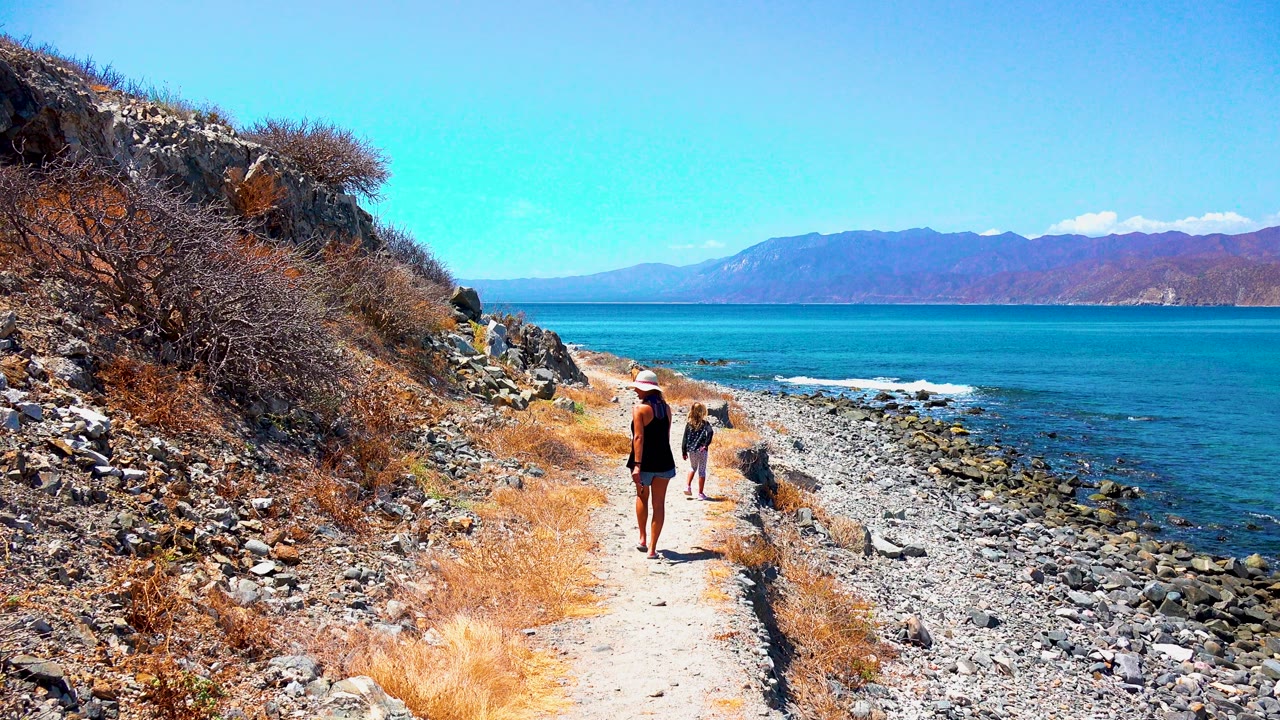 At 0:00 in El Sargento, Mexico, a woman and child walk away on a narrow coastal path with dry brush at left, a rocky shoreline at right, and mountains across the blue water.