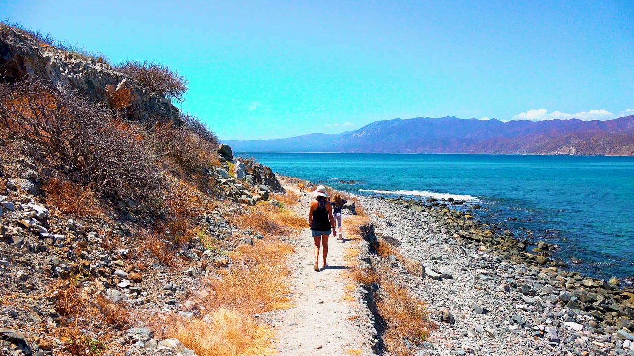 Woman and child continue along a rocky coastal trail under clear sky.