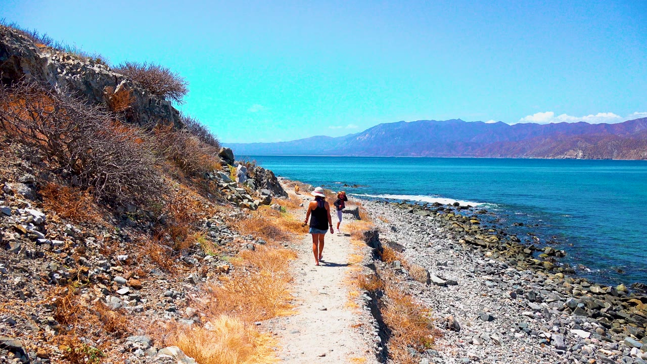 At 0:02 in El Sargento, Mexico, the two figures remain on the trail with the child slightly ahead, dry scrub and loose rocks on the slope, and calm blue water stretching to mountains.
