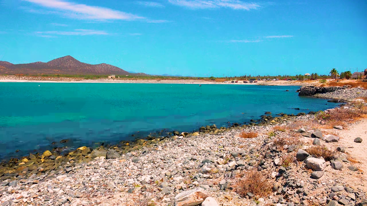 Wide view of the rocky El Sargento La Ventana shoreline with turquoise water, distant mountains, clear blue sky, and no visible people.