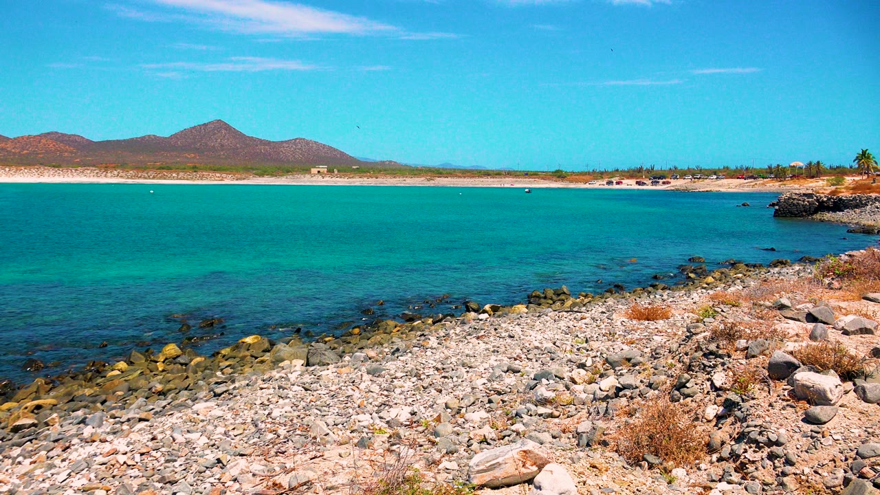 El Sargento La Ventana shoreline one second in, with clear turquoise water across the frame, pebbles in the foreground, distant sand, mountains, and no visible people.