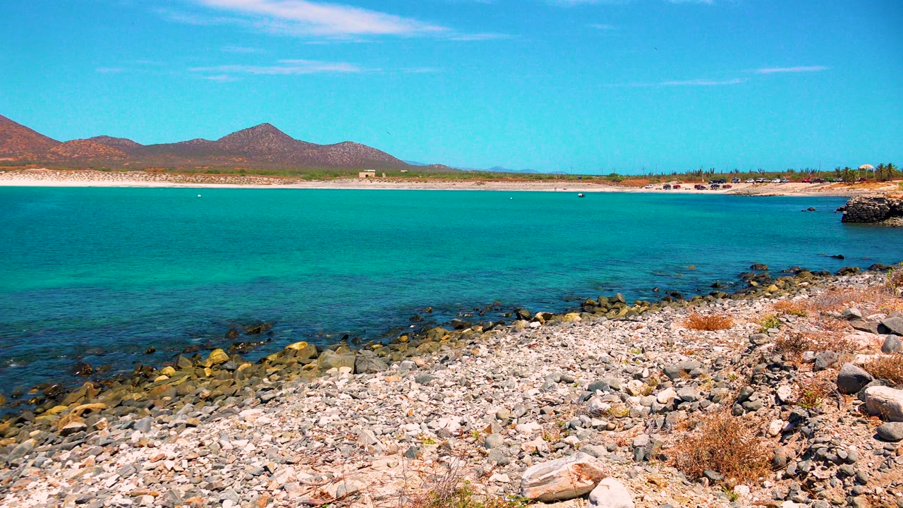 El Sargento La Ventana coastal view two seconds in, with a rocky foreground, clear water shifting near the stones, a pale sandy beach, distant mountains, and no visible people.