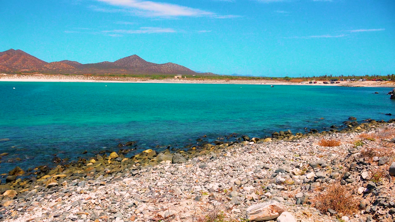 El Sargento La Ventana shore at three seconds, with the rocky foreground lower in frame, calm turquoise sea, a thin sandy beach, mountain ridgeline, and no visible people.