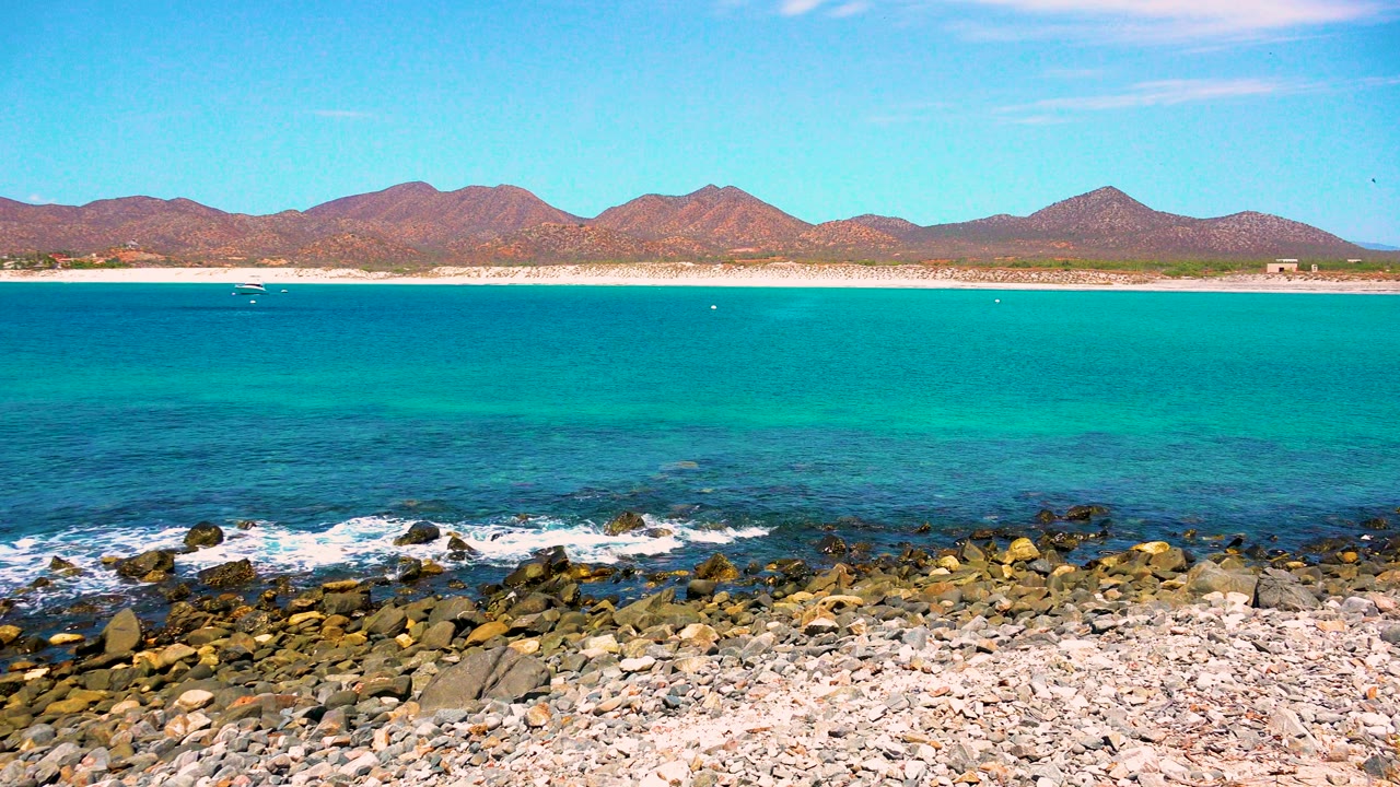 At 0:01 in El Sargento La Ventana, Mexico, a small line of white surf breaks over foreground rocks while the turquoise bay and mountain ridge remain clear, with no visible people.