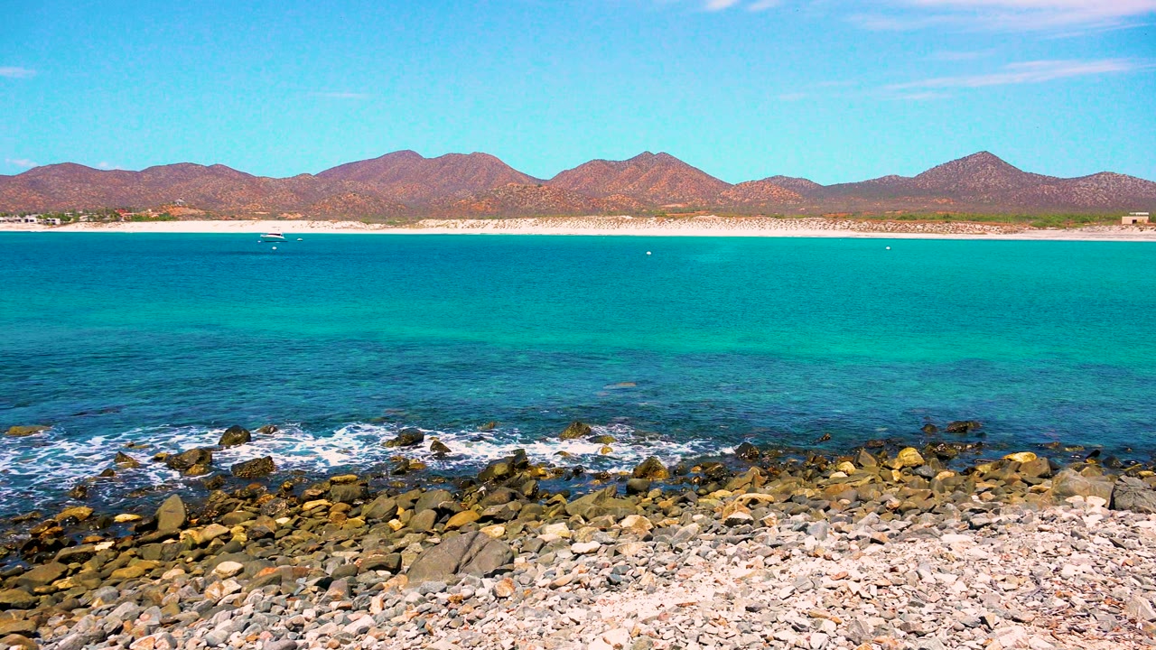 At 0:02 in El Sargento La Ventana, Mexico, the rocky beach foreground sits below smooth turquoise water and a low mountain horizon, with no visible people.
