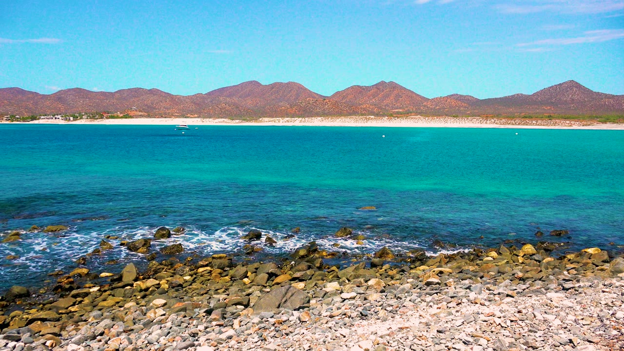 At 0:03 in El Sargento La Ventana, Mexico, blue-green water fills the center of frame above a pebbled shore, with brown mountains across the background and no visible people.