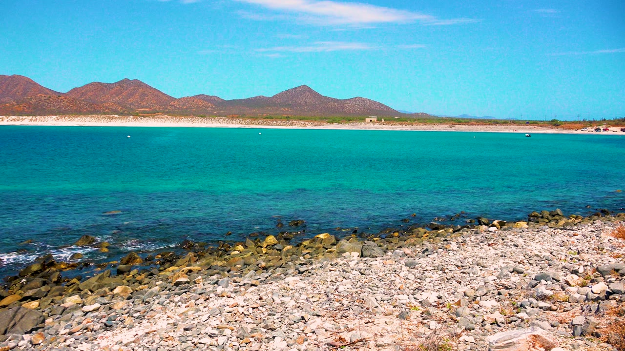Opening frame of El Sargento La Ventana, Mexico with pale pebbles and dark rocks in the foreground, turquoise ocean beyond, distant mountains, and no visible people.