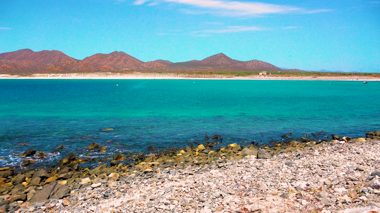Third frame from El Sargento La Ventana, Mexico showing a wider sweep of rocky beach, blue-green water, low mountains across the shore, and no visible people.