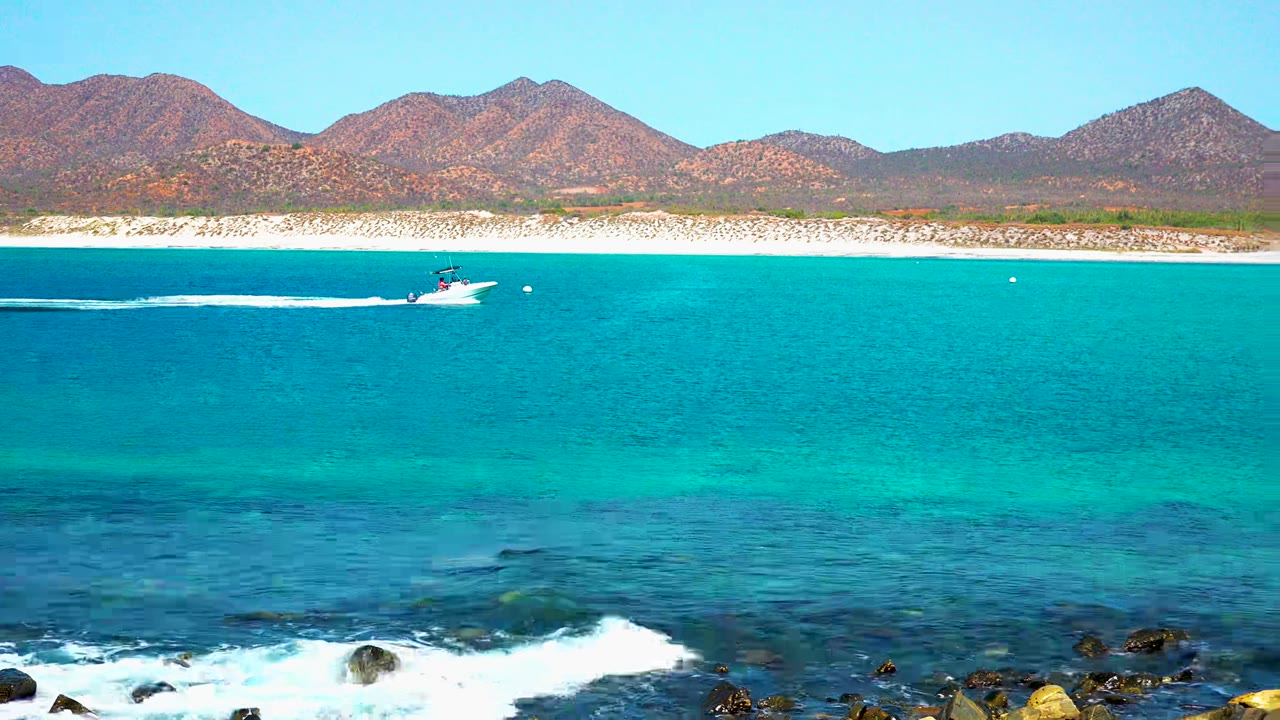 At 0:00, a speedboat crosses turquoise water near El Sargento La Ventana, Mexico, with rocky surf in front, pale beach and mountains behind, and no visible people.