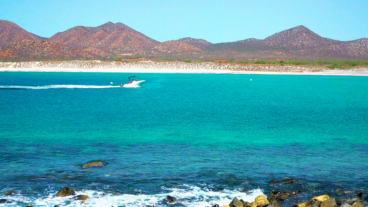 At 0:02, the speedboat continues offshore at El Sargento La Ventana, Mexico, while foamy waves break over dark foreground rocks and no visible people appear.