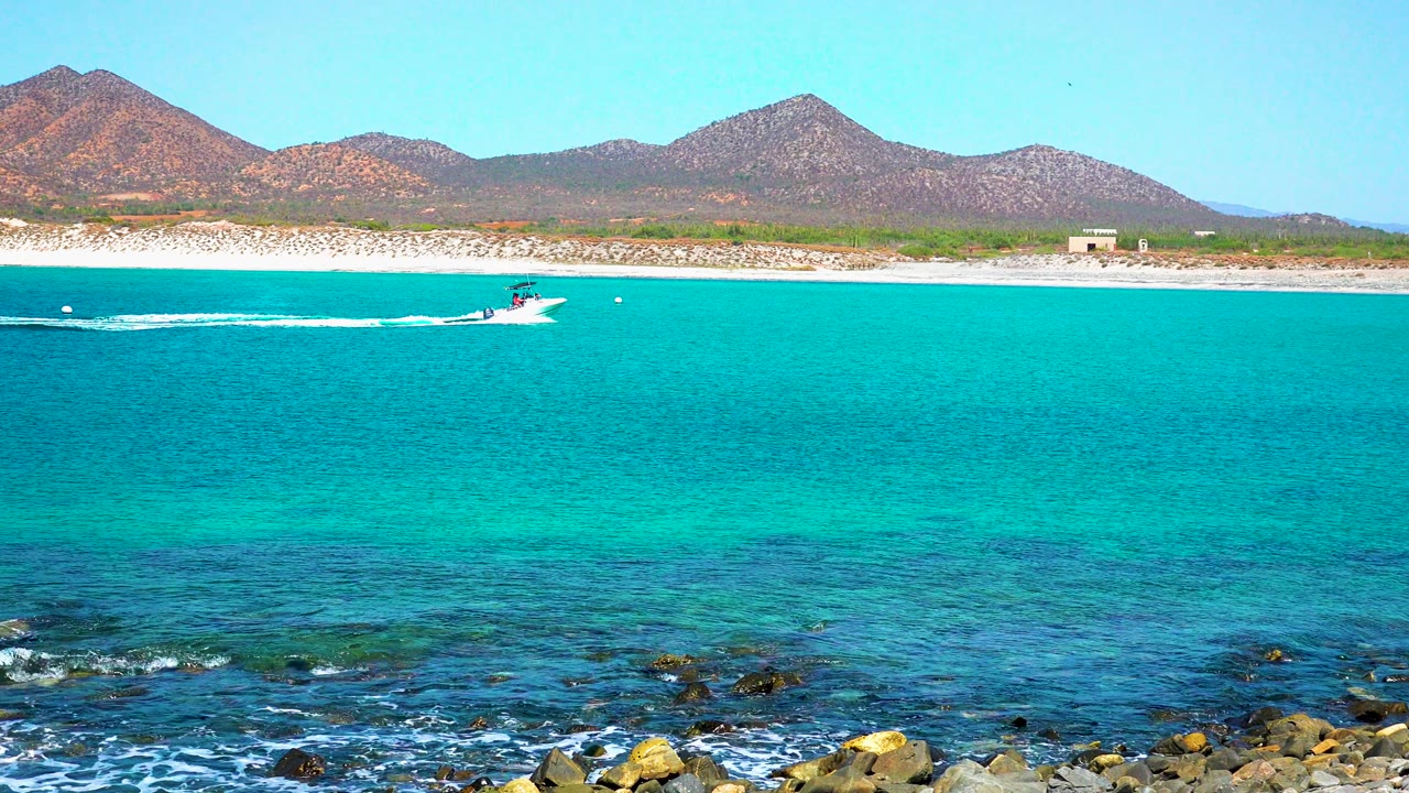 At 0:00 in El Sargento La Ventana, Mexico, a small boat begins crossing turquoise coastal water beyond rocks and foam, with sandy shore, low buildings, and mountains in the background, no visible people.