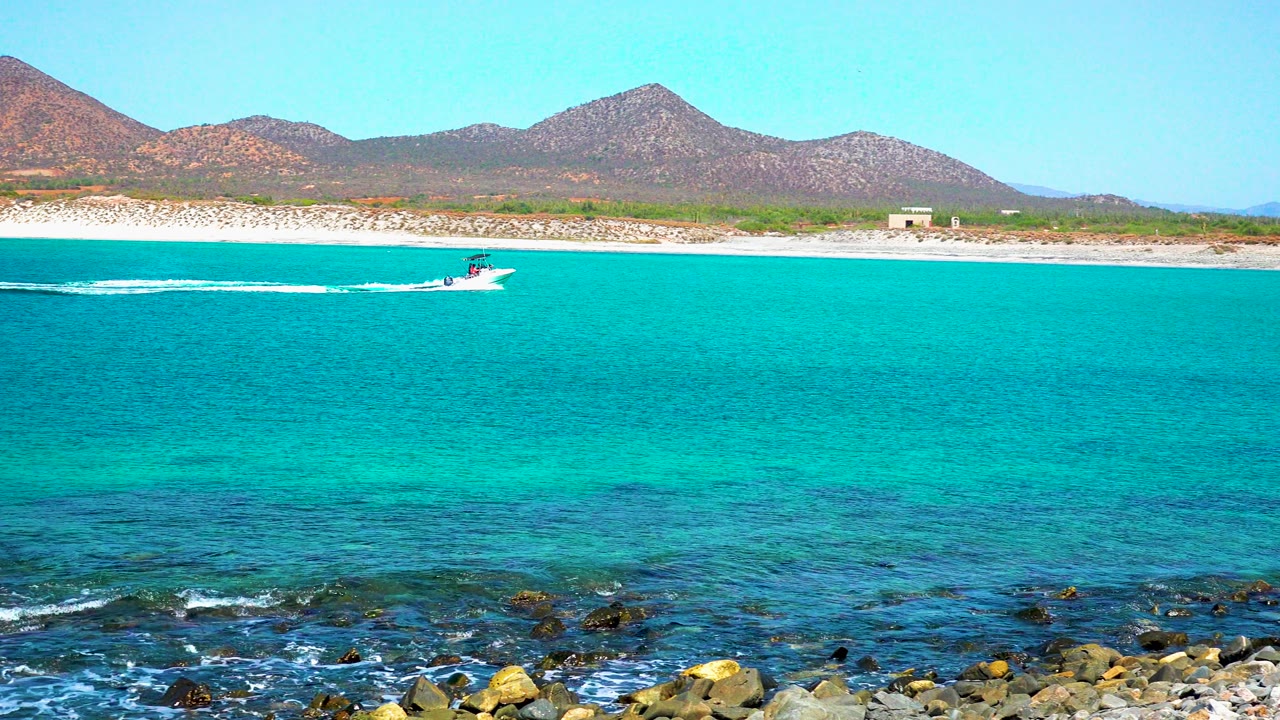 At 0:01 in El Sargento La Ventana, Mexico, the boat has moved farther right across the turquoise water, leaving a thin white wake below the sandy beach and mountain ridge, no visible people.