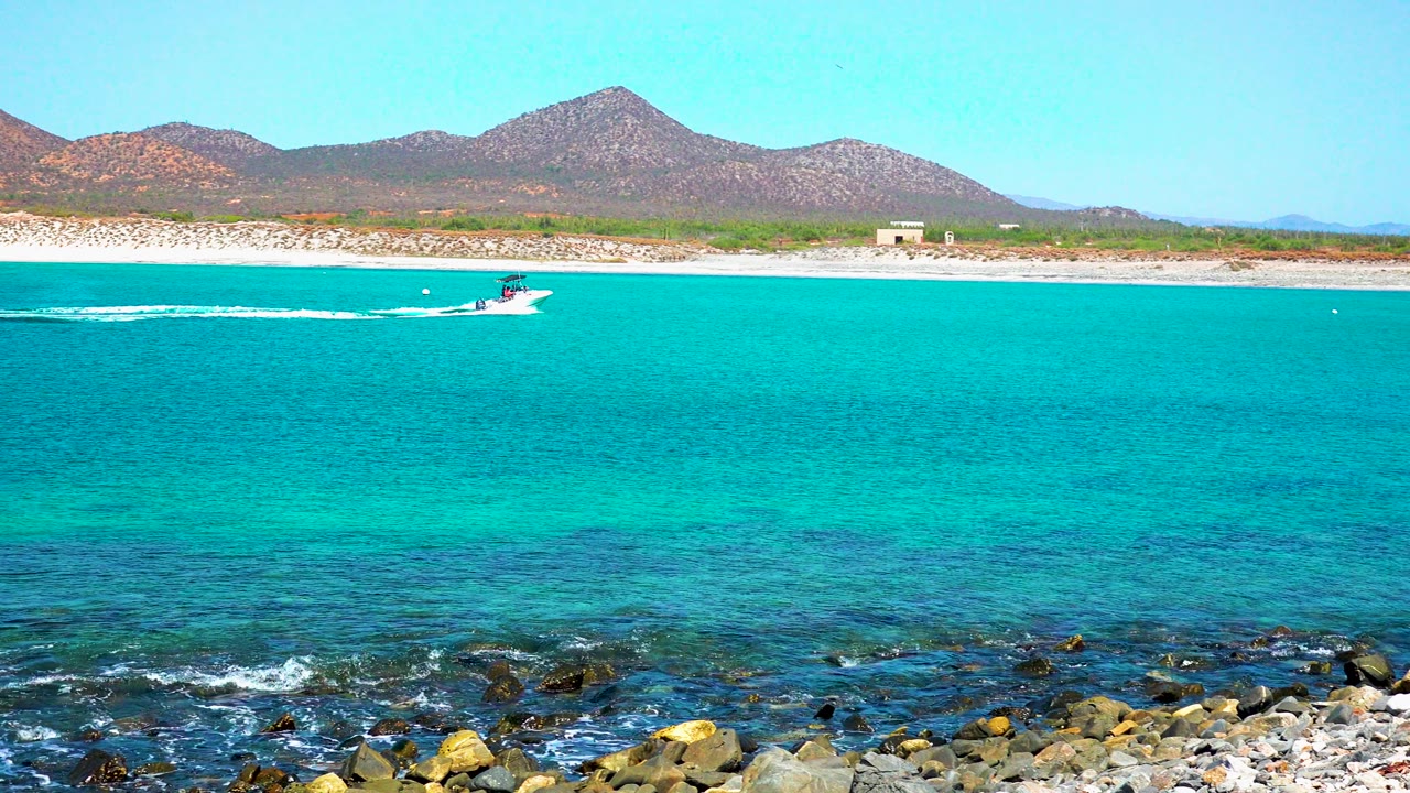 At 0:02 in El Sargento La Ventana, Mexico, the boat continues rightward across bright turquoise water, with darker underwater patches near the rocky foreground and the mountain slope behind the shore, no visible people.