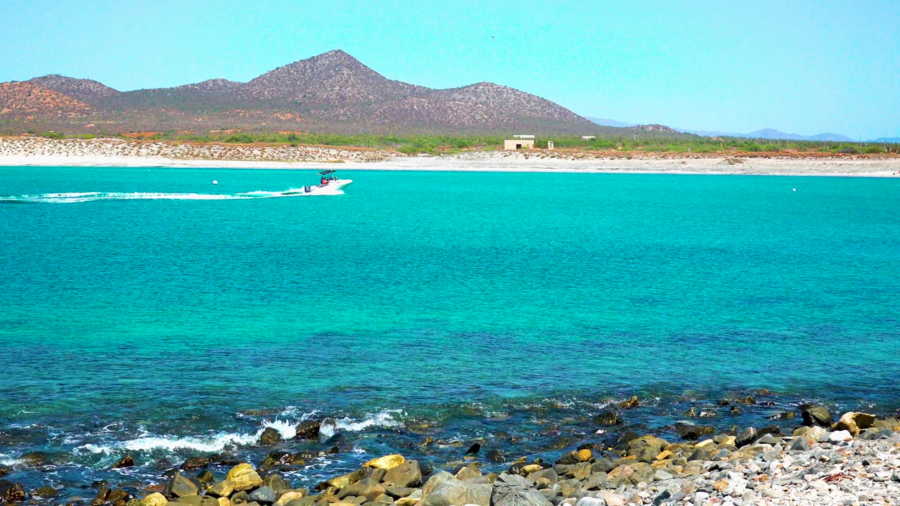 Boat and long wake crossing turquoise water above rocky surf.