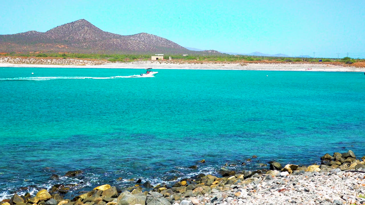 At 0:00 in El Sargento La Ventana, Mexico, a small speedboat crosses turquoise water beyond a rocky foreground shore, with a mountain and clear sky behind it and no visible people.