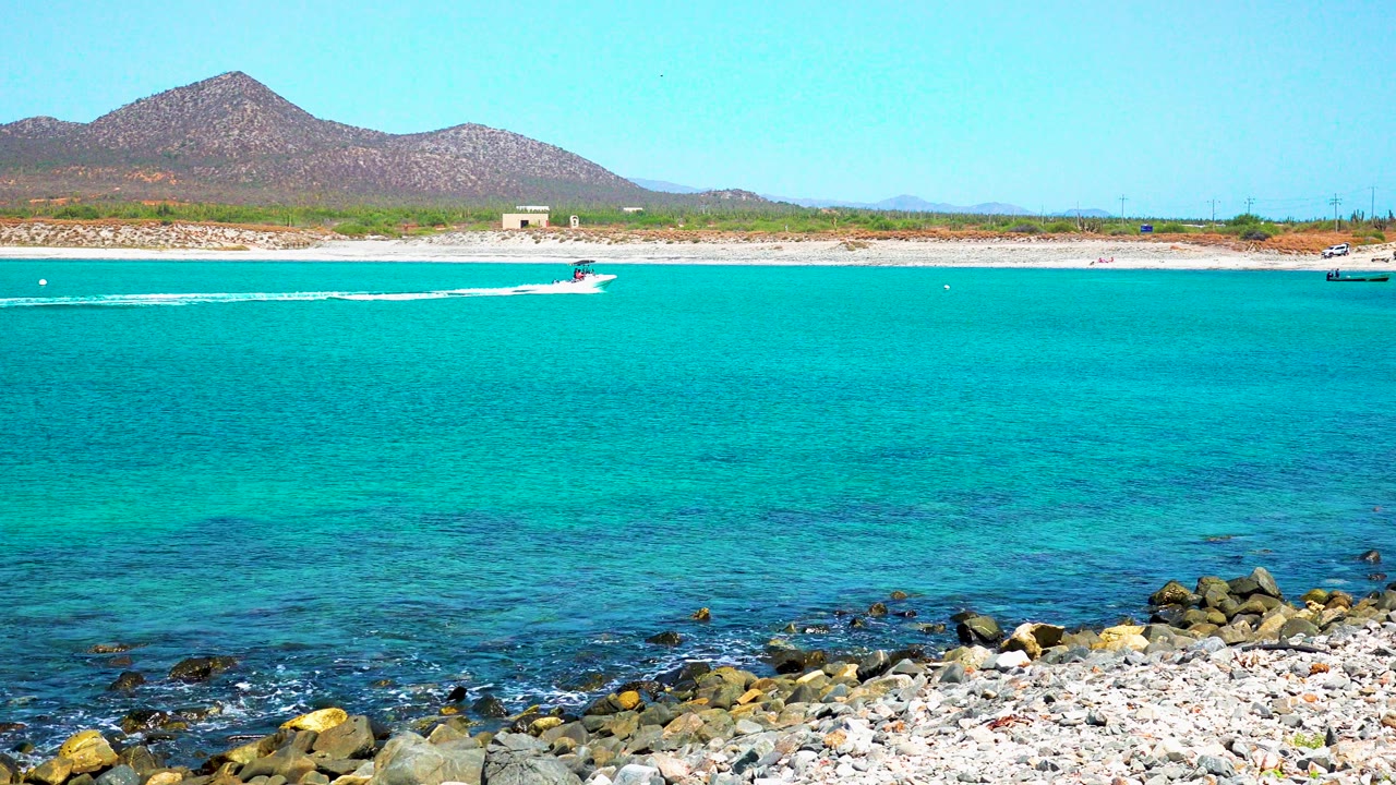 At 0:01 in El Sargento La Ventana, Mexico, the speedboat sits farther across the turquoise bay, trailing a white wake above the rocky beach foreground with no visible people.