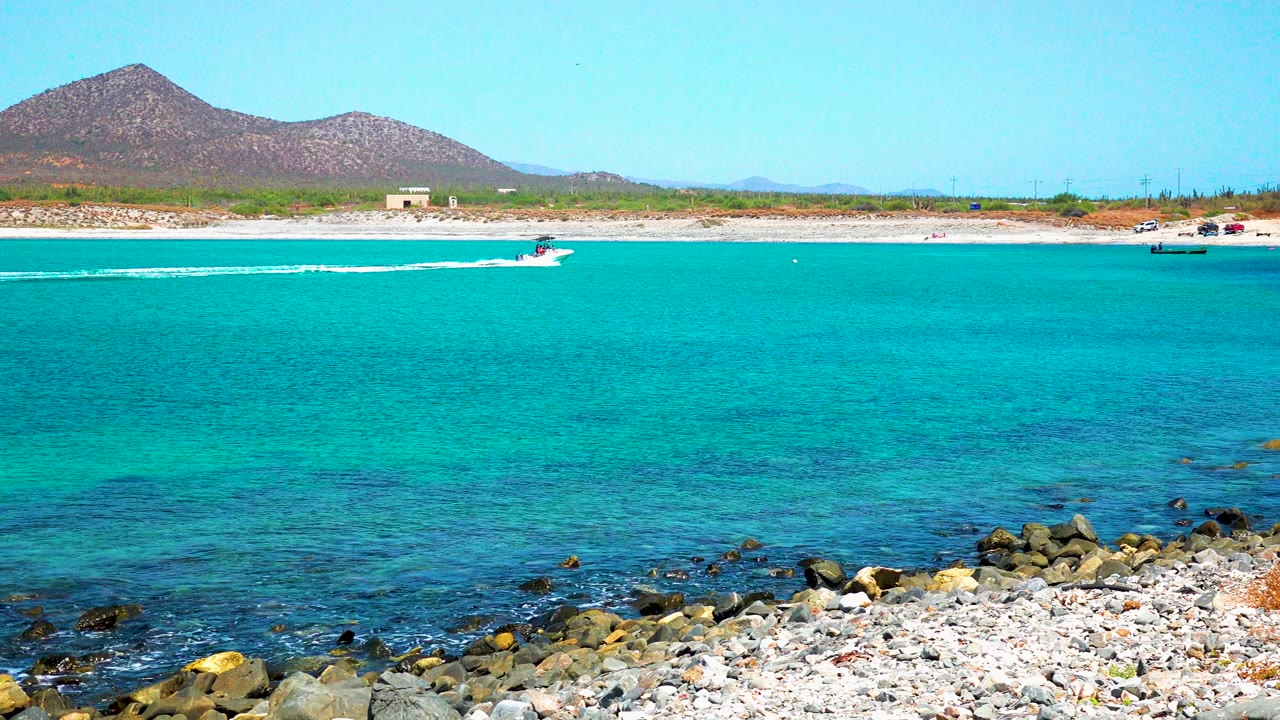 At 0:02 in El Sargento La Ventana, Mexico, the speedboat continues across the turquoise water while distant parked vehicles and a mountain line the beach, with no visible people.